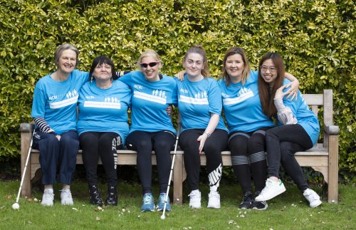 Various NCBI team members in light blue running tops gather around Dr Sinead Kane on a sunny park bench and hold light-hearted stretch poses for the camera.