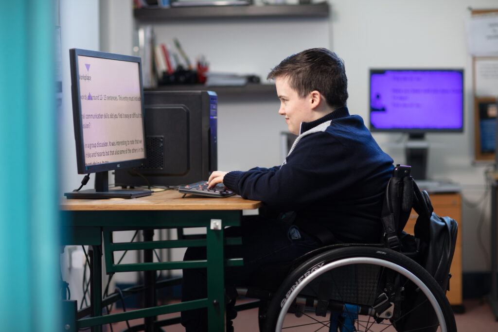 A young boy in a wheelchair using assistive technology and reading large print on a desktop computer