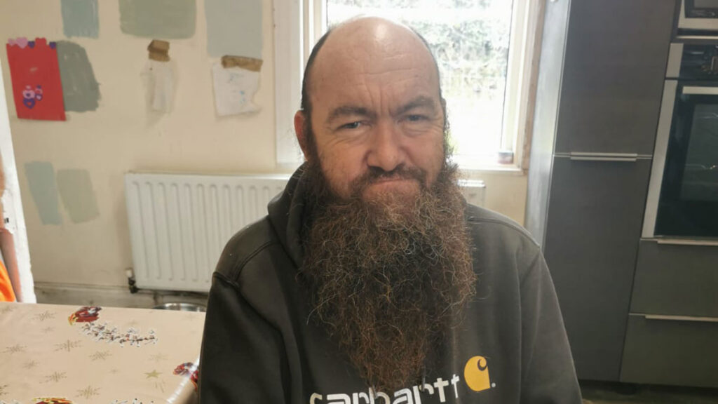 Gerard McHugh who features in this podcast sits on a chair in front of a desk to pose for a picture while he wears a black hoody. Gerard has a long beard.